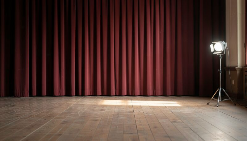 A bare wooden stage floor seen from the wings, a dark red curtain hanging at the edge, a single work light standing near the wall, dust in the beam and no lettering or signs anywhere in view.